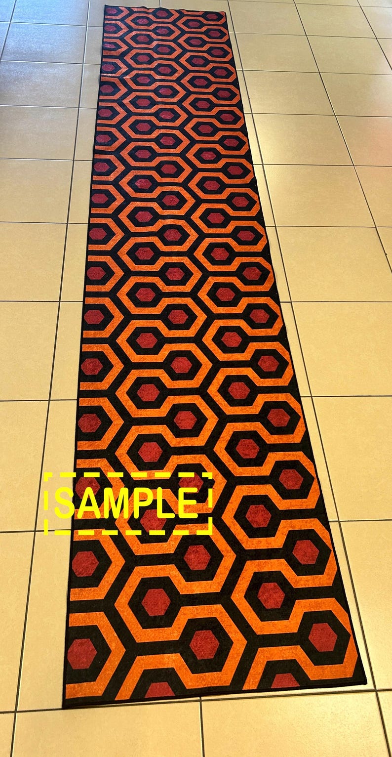 Op de afbeelding: Een lang, rechthoekig tapijt met een herhalend geometrisch patroon. Het ontwerp bevat zwarte, oranje en bordeauxrode vormen. Het woord "SAMPLE" is in het geel zichtbaar. Het tapijt ligt op een betegelde vloer.