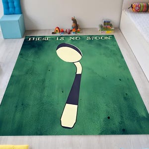 May include: A green rectangular rug featuring a large spoon illustration. The spoon is white, black, and cream-coloured. The text "THERE IS NO SPOON" is printed in white across the top of the rug. The rug is on a light wood floor.