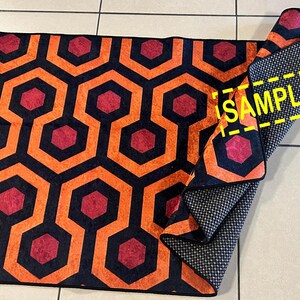 Op de afbeelding: Een opgerolde vloerkleed met een herhalend geometrisch patroon van oranje, zwarte en rode zeshoeken. De onderkant van het vloerkleed is donkergrijs met een klein wit stippenpatroon. Het woord "SAMPLE" is zichtbaar.