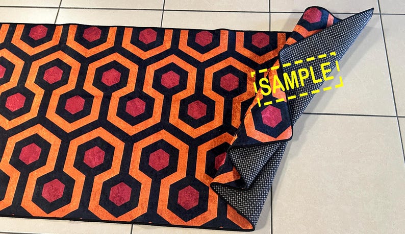 Op de afbeelding: Een opgerolde vloerkleed met een herhalend geometrisch patroon van oranje, zwarte en rode zeshoeken. De onderkant van het vloerkleed is donkergrijs met een klein wit stippenpatroon. Het woord "SAMPLE" is zichtbaar.