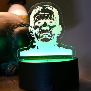 May include: A black and green Frankenstein's monster LED lamp. The lamp features a detailed illustration of the monster's face, illuminated by a green glow. The base is black and round. The lamp is a decorative item.