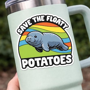 May include: A light green tumbler with a sticker featuring a gray manatee illustration. The sticker has the text "SAVE THE FLOATY POTATOES" in white, with a rainbow background. The tumbler has a silver lid and a handle.
