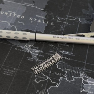 May include: A silver Pentel GraphGear 1000 mechanical pencil with a textured grip and a metal clip, positioned above a small black rectangular label featuring a QR code and the text FAA Registration# FA1234567890 printed in white.