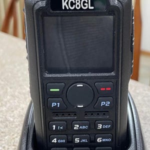 以下が含まれることがあります： 充電ベース付きの黒いトランシーバー。トランシーバーには、画面、ボタン、前面に「KC8GL」というコールサインが印刷されています。充電ベースにも「KC8GL」というコールサインが印刷されています。