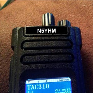 May include: A black handheld radio with a screen displaying "TAC310" and "CH-0013 Zone 003". The radio has a label on the top that reads "N5YHM".