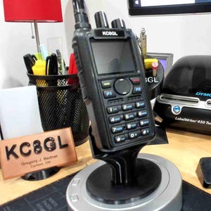 Puede incluir: Una radio bidireccional negra con un teclado y una pequeña pantalla. La radio está sobre una base giratoria negra y plateada. La radio tiene el texto "KC8GL" en la parte delantera. Una placa de identificación con el texto "KC8GL Gregory J. Bednar Canton, OH" está en primer plano.
