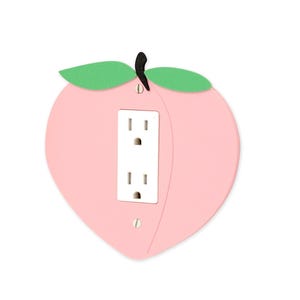 May include: A decorative light pink peach-shaped outlet cover with a white outlet. The peach has green leaves and a black stem. The outlet cover is designed to fit a standard US electrical outlet.