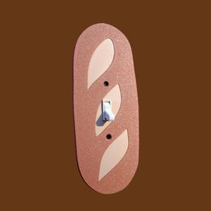 May include: A decorative light switch cover in the shape of a baguette. The cover is a speckled copper color with three off-white leaf-shaped accents and a white toggle switch. The background is a solid brown.
