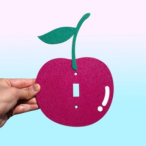 May include: A decorative light switch cover shaped like a cherry. The cover is a vibrant magenta color with a green glittery stem and leaf. The switch opening is rectangular, with two screw holes. The design is held by a hand.