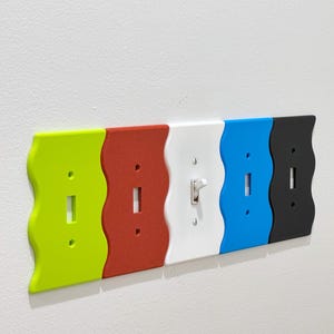 May include: Five light switch covers in different colors: green, red, white, blue, and black. Each cover has a wavy design and a single light switch.