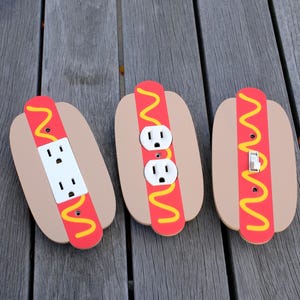 May include: Three decorative hot dog-shaped wall plates. One has a double electrical outlet, another has a single outlet, and the third has a light switch. Each features a tan bun, red hot dog, and yellow mustard design.