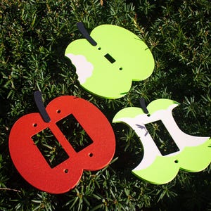 May include: Three apple-shaped light switch covers in red and green, with black stems. Two are green with white and one is red. The green ones have a bite taken out of them. The covers are on a bed of green foliage.