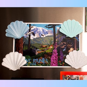 May include: A refrigerator magnet featuring a scenic image of Mount Rainier National Park, with four seashell-shaped magnets in shades of blue and white. The image includes a mountain range, trees, and wildlife.