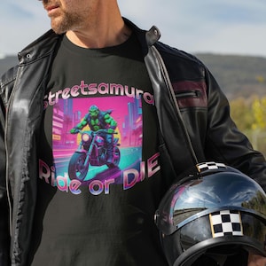 Könnte beinhalten: Ein schwarzes T-Shirt mit einem Aufdruck eines grünen Monsters, das ein Motorrad in einer Neon-Stadtlandschaft fährt. Der Text "streetsamura Ride or DIE" ist auf dem Shirt gedruckt.