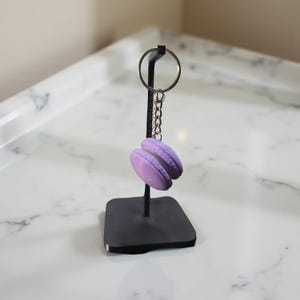 May include: A lavender macaron keychain hangs from a black metal stand. The keychain features two lavender-colored macaron cookies with a white filling. The stand has a square base and a circular key ring.