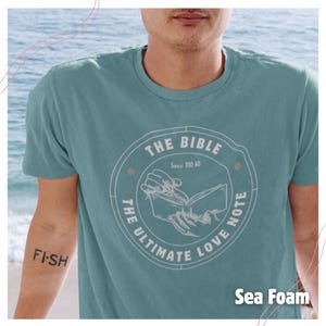 May include: Sea foam green t-shirt featuring a circular design with the words "THE BIBLE" and "THE ULTIMATE LOVE NOTE" around an open book illustration. The text "Since 100 AD" is also included. The word "FISH" is tattooed on the arm.
