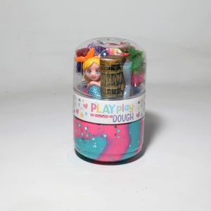 Peut inclure: Un récipient en plastique transparent de Play Play Dough avec une figurine de sirène, une tour miniature et d'autres petits jouets. La pâte est tourbillonnée rose et bleu. Le récipient porte l'inscription "PLAY play DOUGH".