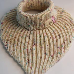 May include: A cream-coloured knit cowl neck warmer featuring a vertical ribbed texture and multicoloured flecks of blue, pink, and green throughout the yarn. A small pink rectangular tag is attached to the side of the ribbed collar.