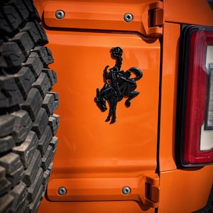 Könnte beinhalten: Ein glänzend schwarzes Emblem, das einen bockenden Bronco mit Reiter darstellt, angebracht auf der leuchtend orangefarbenen Rückwand eines Fahrzeugs. Das Emblem befindet sich zwischen einem großen schwarzen Reifen links und einem rot-weißen Rücklicht rechts.