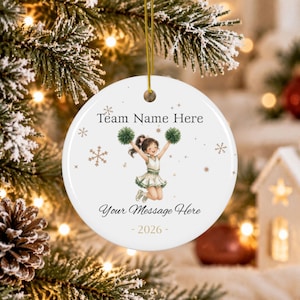 Può includere: Un ornamento rotondo in ceramica bianca con un cordino dorato, un'illustrazione centrale di una cheerleader che salta con i pon pon e il testo Team Name Here, Your Message Here e 2026, circondato da piccoli fiocchi di neve dorati.