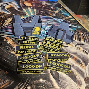 Op de afbeelding: Een set 3D-geprinte spelstukken voor een bordspel. De stukken zijn zwart met gele tekst en bevatten het volgende: +1 SEC RUSH, RUSH, DP PROT, -3000 DP, -1000 DP, -4000 DP, BLOCKER, REBOOT, REBOOT, ALLIAN, +2000 DP, +3000 DP, +2000 DP.