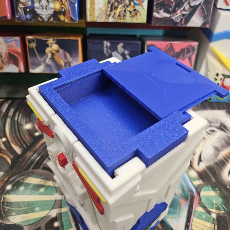 May include: A white, blue, red, and yellow plastic storage container with a hinged blue lid. The container features a rectangular opening at the top, a textured surface, and geometric moulded details on the exterior sides and front panel.