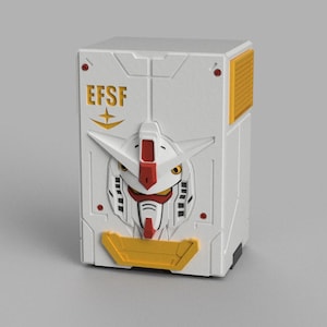 Puede incluir: Una caja blanca y amarilla con un diseño de rostro de robot estilizado. La caja tiene las letras "EFSF" en dorado. El rostro del robot tiene detalles rojos, blancos y negros. La caja es rectangular con un panel amarillo en el lateral.