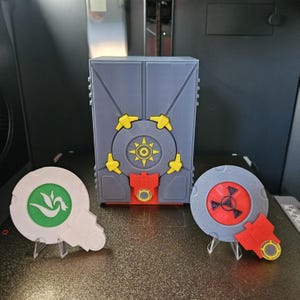 Può includere: Una scatola grigia, gialla e rossa stampata in 3D con un design circolare e due accessori circolari abbinati. Un accessorio è bianco con un design verde, l'altro è grigio con un design rosso.