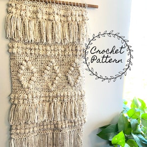 May include: A beige crochet wall hanging with a textured pattern and fringe. The hanging is attached to a wooden dowel rod and is displayed on a white wall. The text "Crochet Pattern" is written in a circle with leaves around it.