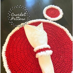 May include: Red crochet placemat with a white crocheted border. A white napkin is placed on top of the placemat with a white crocheted napkin ring. A smaller red crochet coaster with a white crocheted border is in the background.