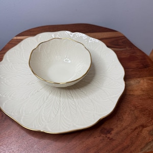 May include: A cream-colored, leaf-patterned serving dish set. The set includes a large, scalloped-edge plate and a smaller bowl, both with gold trim. The bowl sits atop the plate, creating a layered presentation. The set is designed for serving food.