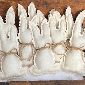 May include: A group of eight handmade white fabric bunny rabbits with brown twine tied around their necks. The bunnies are arranged in a row on a wooden surface.