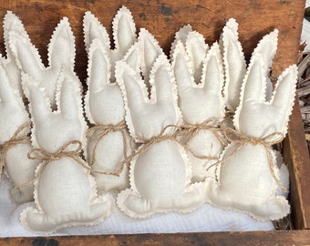 May include: A group of eight handmade white fabric bunny rabbits with brown twine tied around their necks. The bunnies are arranged in a row on a wooden surface.