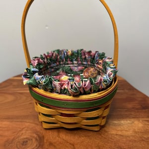 May include: A small, woven basket with a yellow base and a tall, arched handle. The rim is decorated with a green and red stripe, and the interior features a floral fabric lining. The basket is suitable for Easter or spring decor.