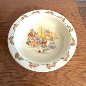 May include: A white ceramic bowl with a border of brown bunnies hopping around the edge. The center of the bowl features a colorful illustration of four bunnies gathered around a television set.