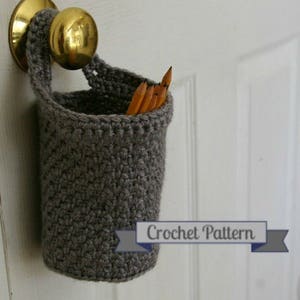 May include: A grey crocheted pencil holder hanging on a door knob. The holder is filled with pencils. The text "Crochet Pattern" is visible below the holder.