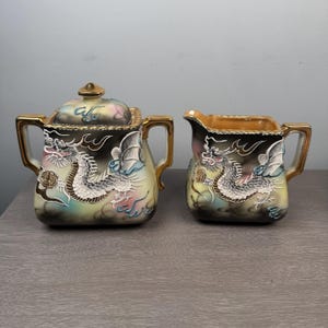 May include: A ceramic sugar bowl with a lid and a matching creamer pitcher. Both pieces feature a black and cream gradient background with a raised, white, grey, and blue dragon motif, gold-painted handles, and gold-trimmed edges on the rims.