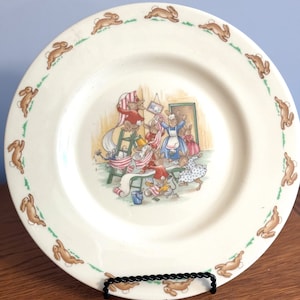 May include: A white ceramic plate with a green border and a repeating pattern of brown bunnies hopping around the edge. The center of the plate features a colorful illustration of a group of bunnies decorating a room with a banner.