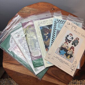 May include: A collection of sewing patterns in clear plastic sleeves. The patterns feature illustrations of dolls and other crafts. Some patterns have text that reads "SEWN FROM THE" and "Down Emory Lane". The patterns are on a wooden surface.