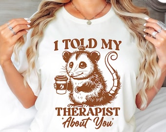 May include: Natural-colored t-shirt with a brown graphic of an opossum holding a coffee cup. The text reads "I TOLD MY THERAPIST About You". The shirt is made of a soft material.