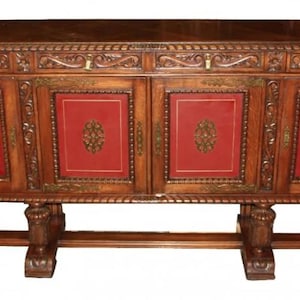 May include: A large, antique wooden sideboard with four doors and two drawers. The doors are carved with intricate details and have red leather panels. The sideboard is supported by four carved legs.