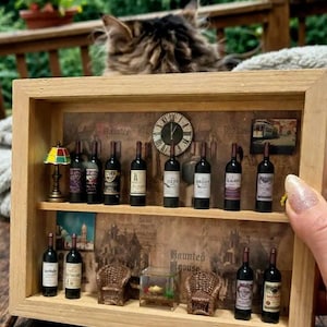 May include: A miniature wooden display case with two shelves, showcasing tiny wine bottles, a lamp, a clock, and miniature furniture. The background features a vintage-style print with the words "Haunted House". A cat is visible in the background.
