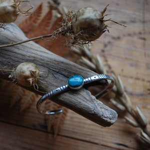 Peut inclure: Un bracelet en argent avec une pierre turquoise sertie au centre. Le bracelet est posé sur un morceau de bois flotté avec des fleurs séchées.