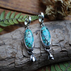 May include: A pair of silver earrings with oval turquoise stones. The stones have a mottled pattern of blue and black. Each earring has a small silver flower charm at the bottom. The earrings are displayed on a piece of driftwood.