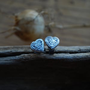 May include: Two silver heart-shaped earrings with intricate floral designs. The earrings are on a wooden surface.