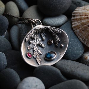 Peut inclure: Un collier pendentif en argent avec un motif de feuille et trois petites pierres précieuses : rouge, marron et bleu. Le pendentif est posé sur un fond de pierres grises et un coquillage.