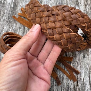 May include: A brown leather belt featuring a thick, braided construction with multiple long, flat leather fringe strips hanging from the ends. The leather has a matte, textured finish with visible grain and natural variations in the brown color.
