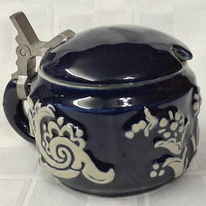 May include: A dark blue ceramic beer stein with a hinged metal lid and handle. The stein features white floral and scrollwork designs around the body. The lid has a small, curved handle. The stein is likely used for drinking beer.
