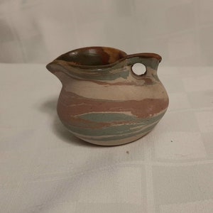 May include: A small, handcrafted ceramic pitcher with a unique design. The pitcher features a spout and a handle with a hole. The body is adorned with horizontal bands of muted colors, including pink, green, and beige. The rim is a darker brown.