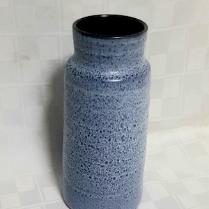 May include: A cylindrical ceramic vase featuring a speckled light blue and dark blue glaze. The vase has a slightly narrower neck and a smooth, dark interior rim. The exterior surface has a textured, mottled appearance with a consistent color pattern.